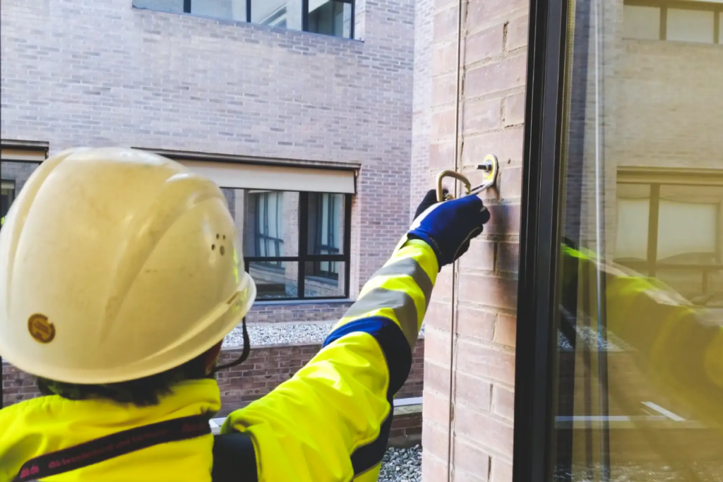 Vista de una trabajador, vestido con EPIs (casco, guantes de seguridad y chaqueta de alta visibilidad) a punto de sujetarse a un anclaje estructural en la cornisa de un edificio.