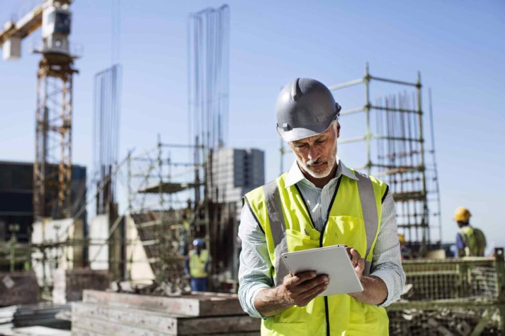 Imagen en primer plano de una persona, el Coordinador de Seguridad de una obra, con chaleco reflectante y casco, con una tablet en la mano, delante de una obra de construcción.